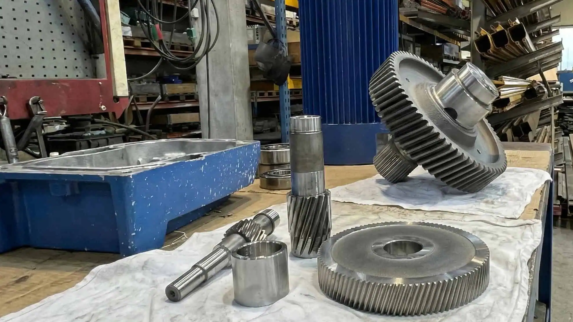 Flere store metalgear og -aksler er anbragt på hvide klude på en arbejdsbænk i et industrielt værksted, hvor man kan se hylder og udstyr i baggrunden.