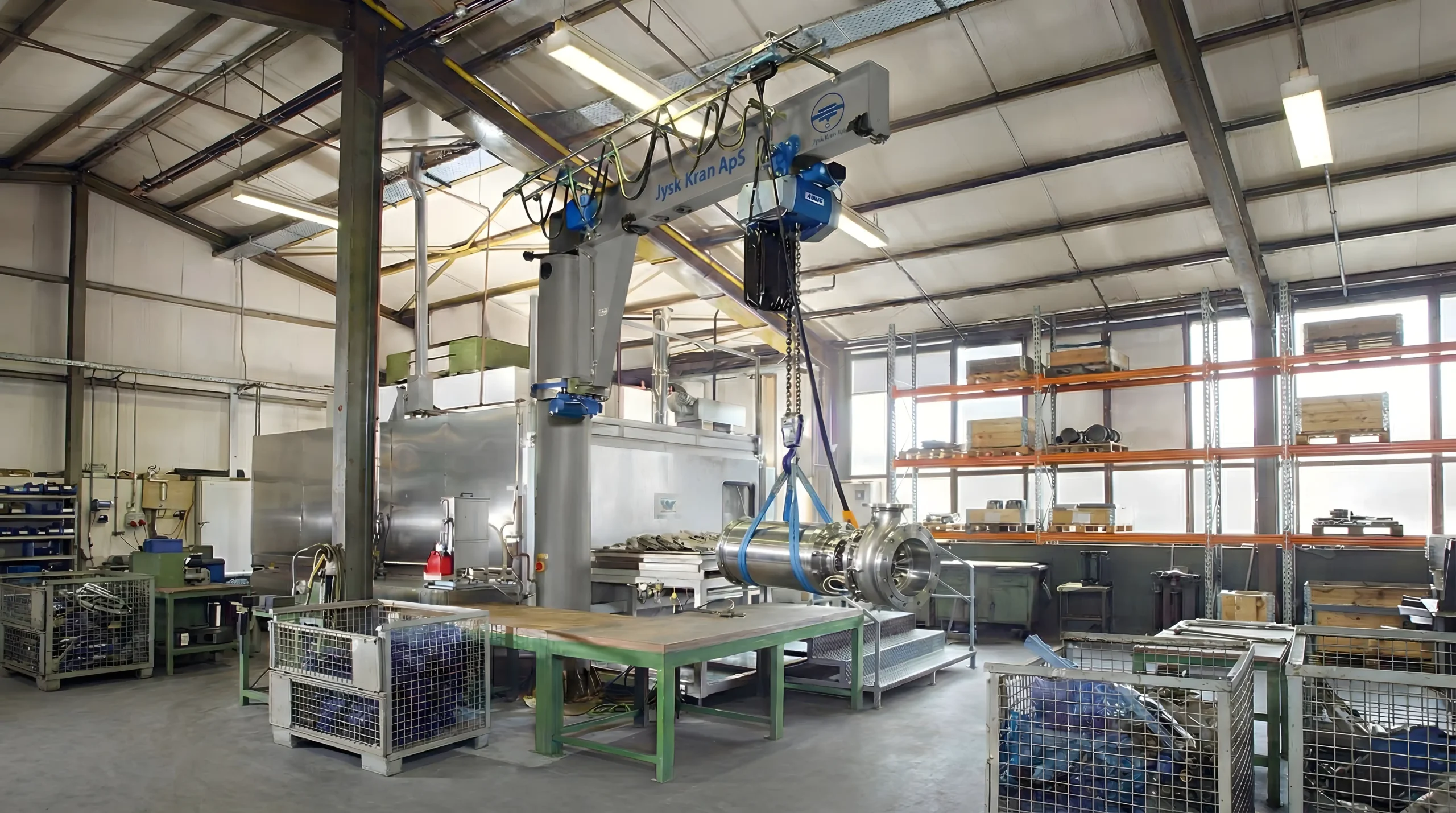 Et rummeligt industrielt værksted med en stor robotarm, der løfter en cylindrisk metalgenstand over et arbejdsbord. Hylder, metalkasser og forskelligt værktøj og udstyr er organiseret rundt omkring i lokalet.