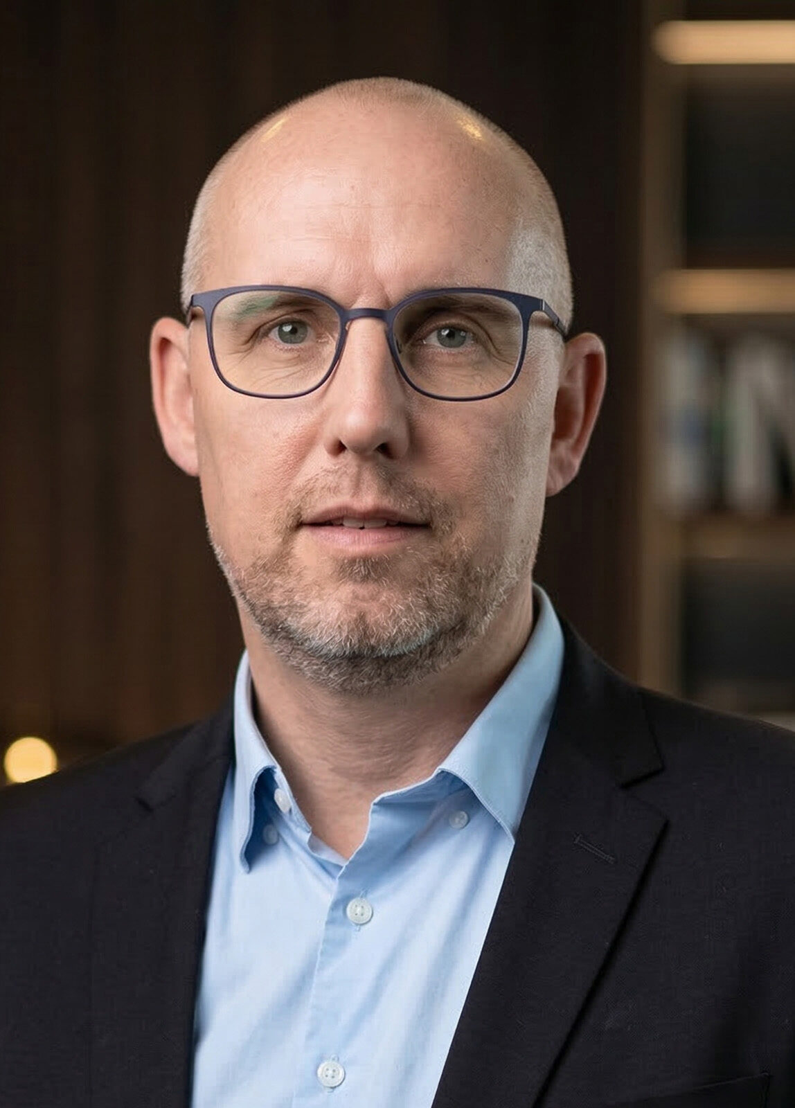 En mand med kort hår, briller og trimmet skæg iført lyseblå skjorte og mørk habitjakke står foran en sløret baggrund med hylder og varm belysning.